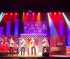 A man in a red jacket and a woman in a sparkly dress sit across from each other at a table on stage, with the man holding a microphone and a 