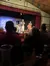 The image shows an audience watching performers on a stage within a dimly lit, intimate theater setting.