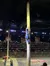 A performer is climbing a tall pole in front of an audience while others watch from the ground during what seems to be a lumberjack show or similar event.