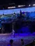The image depicts a dimly-lit theatrical scene with performers dressed in Native American-inspired costumes on a stage that resembles a rugged landscape, with teepees projected in the background.