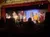 The image depicts a live stage performance with actors in costume engaging with an audience at a theater.