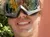 Three individuals wearing ski goggles are posing for a selfie in a sunny, forested area, with the camera holder's finger partly obscuring the lens.