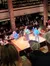 The image shows a lively scene of people watching a performance where two individuals appear to be on stage in a venue with multiple levels of audience seating.