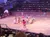 A group of performers dressed in traditional Western attire are engaging in a theatrical scene in a sand-covered arena, while a large audience watches from surrounding tiered seating.