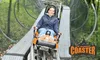 A person is riding a single-person coaster on tracks through a forested area, and the image includes the logo