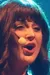 A person dressed in a bright red top and beige fringe skirt is performing on stage with a microphone headset.