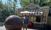 A man stands beside a large spherical fountain near an ice cream concession stand on a sunny day.