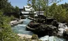 The image depicts a serene artificial river with cascading waters flanked by rocky banks, lush greenery, and rustic structures, possibly a part of an amusement park or themed attraction.