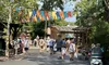The image displays a bustling theme park pathway lined with trees and festooned with colorful banners, with visitors strolling, some pushing strollers, under the bright and sunny sky.