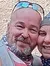 Two cheerful individuals are posing for a selfie in a forested area on a dirt trail.
