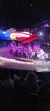 Riders on horseback carry striped flags in a patriotic performance, illuminated under theatrical lighting with an audience in the foreground.