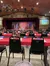 The image shows a dinner theater setup with numbered tables, black chairs, red tablecloths, and a stage with a rustic set design and lighting equipment overhead.