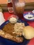 A hearty meal consisting of a grilled pork rib, shredded meat, baked beans, coleslaw, a bread roll, alongside a bowl of cream soup and a glass of iced tea, with condiments like ketchup and mustard present on the table.