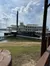 The image presents a large, multi-deck riverboat with iconic paddlewheel elements, docked beside a grassy waterfront under a partly cloudy sky.
