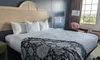 The image shows a neatly arranged hotel room with a large bed, side lamps, and a window overlooking a scenic outdoor view.