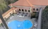 The image shows a bird's-eye view of a kidney-shaped swimming pool with a few people in it, surrounded by lounge chairs and a hotel-like building with an open patio area, taken from behind a window or barrier.