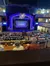 The image shows an audience seated at tables inside a venue, looking towards a stage with a decorative arch that reads