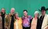 Six people are standing together smiling for a photo in front of a green backdrop, with two men wearing cowboy hats on the far sides and their diverse attire ranging from casual to slightly dressy.