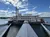 The image displays a large, multi-deck riverboat with a traditional paddlewheel, docked at a pier with a walkway leading up to it under a partly cloudy sky.