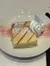 A slice of cheesecake with a dollop of whipped cream is served on a white plate, garnished with powdered sugar and a drizzle of strawberry sauce.