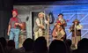 The image shows a group of five performers on stage in front of an audience, dressed in a variety of colorful and thematic costumes that suggest a period or comedic play, with a rustic backdrop setting the scene.