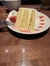 A slice of layered vanilla cake with white frosting, garnished with a strawberry and decorative sauce, is served on a white plate with a fork on the side, accompanied by a cup and glass on a wooden table.