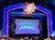 The image shows a brightly lit stage with ornate decorations and the words
