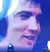 An Elvis impersonator performs on stage with a band, while a large screen displays a close-up of someone's face, and the audience watches and captures the moment.