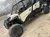 This image shows a side view of a cream-colored Can-Am Commander side-by-side utility task vehicle (UTV), parked on a dusty ground with trees and other vehicles faded in the background.