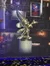 This image features a metallic statue of an animated character with a pointed hat, gesturing towards the viewer, placed on a pedestal within a room filled with shelves of various objects, under ambient lighting with a portrait of a character in a red cloak in the background.