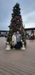 A couple stands happily in front of a large, decorated Christmas tree on a wooden deck with festive polar bear statues.