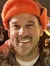 Three people are smiling and posing together, wearing festive holiday accessories like reindeer antlers, an orange hat, and Christmas lights.