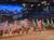 The image shows a live indoor performance with horseback riders holding flags, set in an arena with an audience in bleachers, and a backdrop depicting a snowy mountain scene.