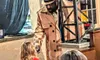 A person in a trench coat is interacting with a child in a dress, while another woman observes them in a restaurant setting.