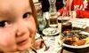 A child is smiling at the camera while sitting at a restaurant table with various dishes, including a dessert, in front of them.