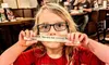 A child wearing glasses and a nametag holds a slip of paper over their mouth in a restaurant setting.