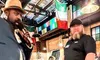 A bearded man in a hat appears to be speaking or singing into a microphone in a lively restaurant setting with colorful flags and patrons in the background.