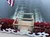 The image shows the stern of a paddlewheel steamboat with red paddlewheels churning water as it moves forward, accompanied by a waving flag.