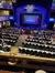The image shows an auditorium filled with people seated at tables, facing a stage with