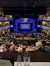 The image shows an audience seated at tables in a theater setting, enjoying a meal while watching a show called