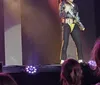 A performer in a glittery outfit sings energetically on stage under dramatic lighting, with an audience watching from below.
