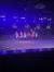 A group of performers on horseback, holding American flags, are showcasing in an arena with an audience watching under blue stage lighting.