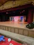The image shows a stage set with a rustic barn theme, complete with wooden barrels, a barn door, and red curtains, under warm stage lighting.