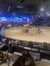 The image depicts a live performance in a themed arena setting, featuring horses and covered wagons with an audience watching from their seats.