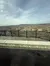 The image shows a scenic landscape with a wrought iron fence in the foreground and a vast view of a town and hills under a partly cloudy sky.