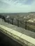The image shows a scenic view of a winding river, a town, and wooded areas in the distance, seen from behind a black metal fence atop a stone wall.