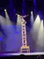 A performer balances on a tall stack of chairs in a spotlight on stage.