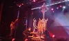 A performer is on a stage suspended at the top of a vertical spinning contraption, surrounded by dramatic lighting and technical staff in action.