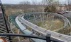 The image shows a winding track, likely for a roller coaster or alpine coaster, surrounded by trees and a parking area with several cars visible.