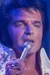 A performer is singing on stage, dressed in an embellished white jumpsuit with a high collar.